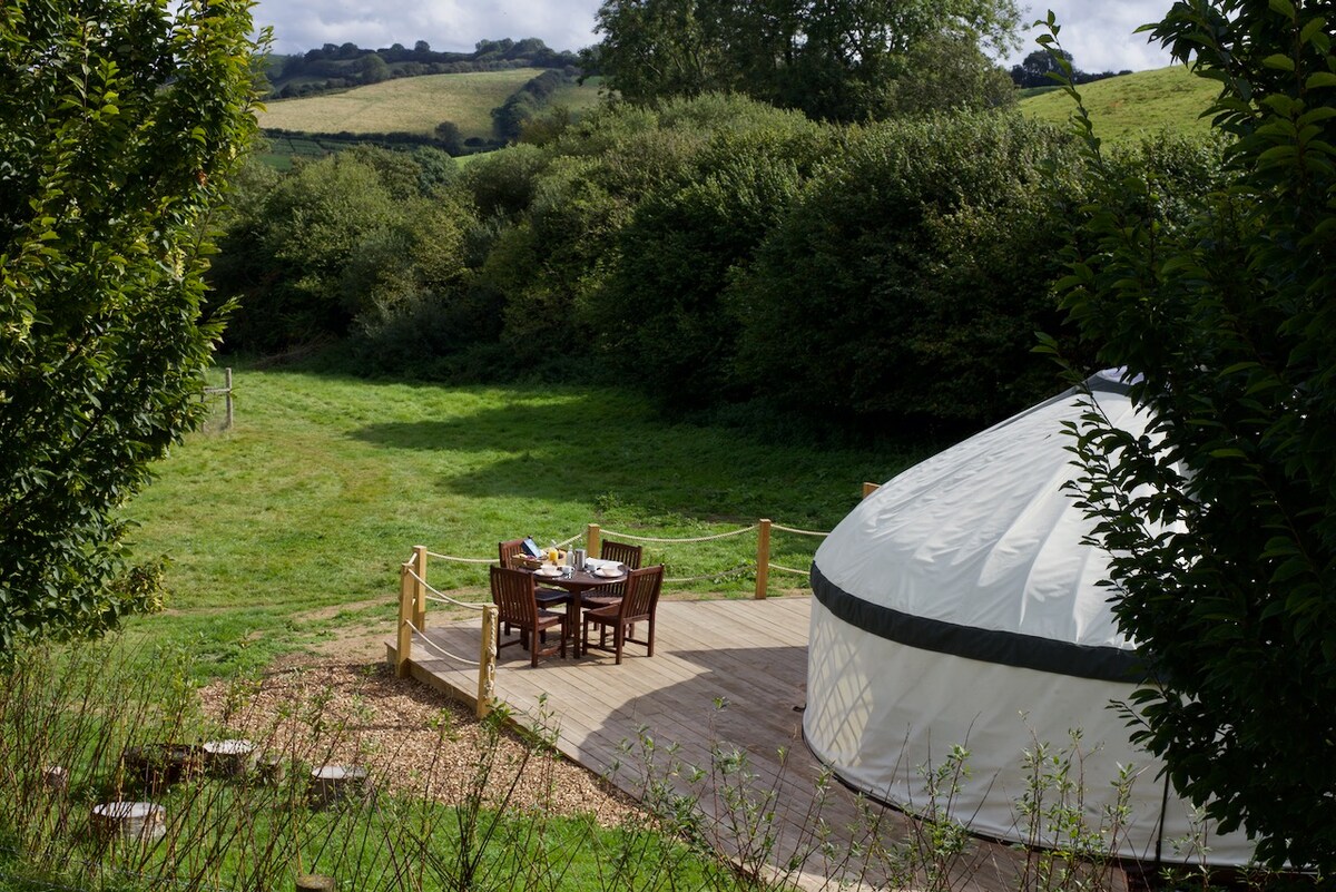A yurt sits on a wooden deck, surrounded by lush greenery and rolling hills. A dining table is set for four, showcasing a serene outdoor space. The clear sky overhead offers a glimpse of the expansive natural landscape.