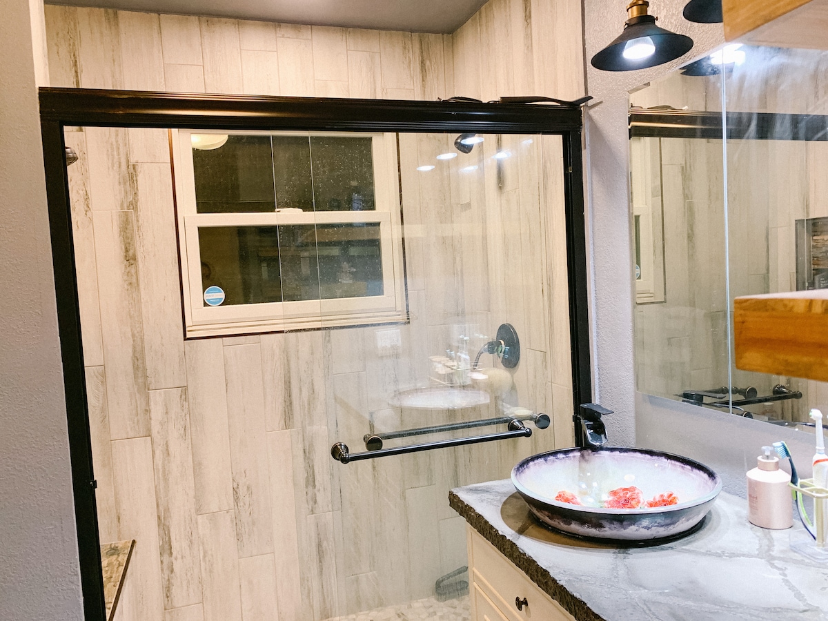 A modern bathroom is showcased, featuring a glass shower enclosure that allows for natural light from a small window. The sleek countertop is complemented by a round vessel sink adorned with decorative elements. Subtle lighting accents the overall design.