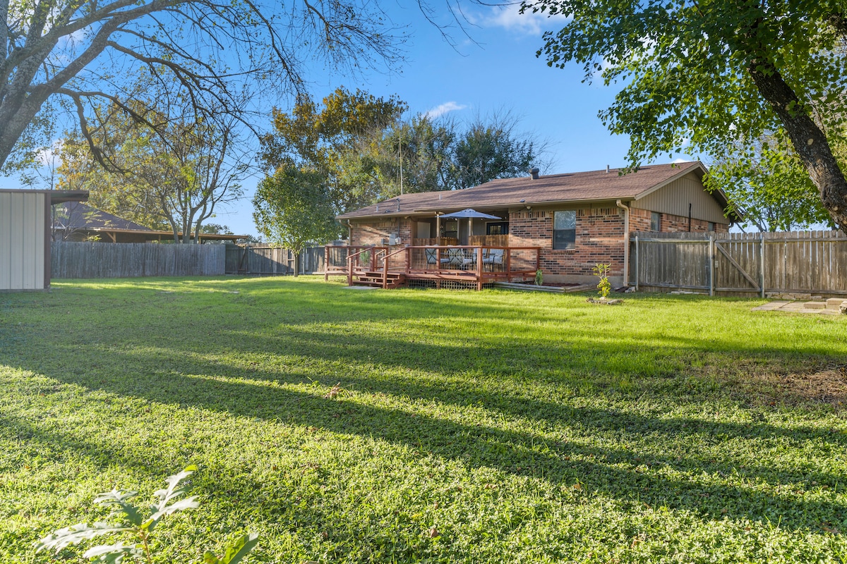 A spacious backyard is shown, featuring well-maintained green grass and a wooden deck attached to the house. The area is surrounded by a wooden fence and several trees, providing shade and a feeling of seclusion.