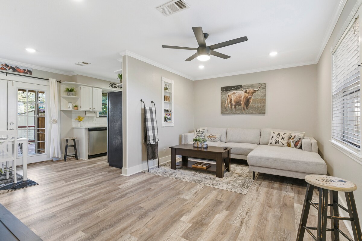 A cozy living area features a light-colored sectional sofa facing a coffee table. A large cow print artwork adorns the wall, while a ceiling fan provides comfort. The space connects to a fully equipped kitchen, with natural light streaming through a nearby door.