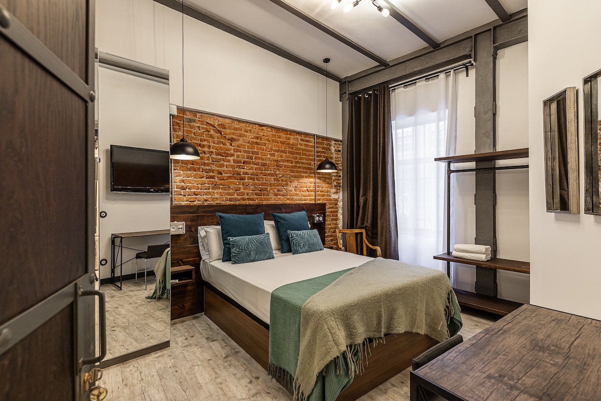 A bedroom is presented with a spacious bed dressed in light linens, complemented by textured throw pillows. Exposed brick walls add warmth, while modern lighting fixtures enhance the space. A small desk, television, and storage shelves are visible, providing functional elements for guests.