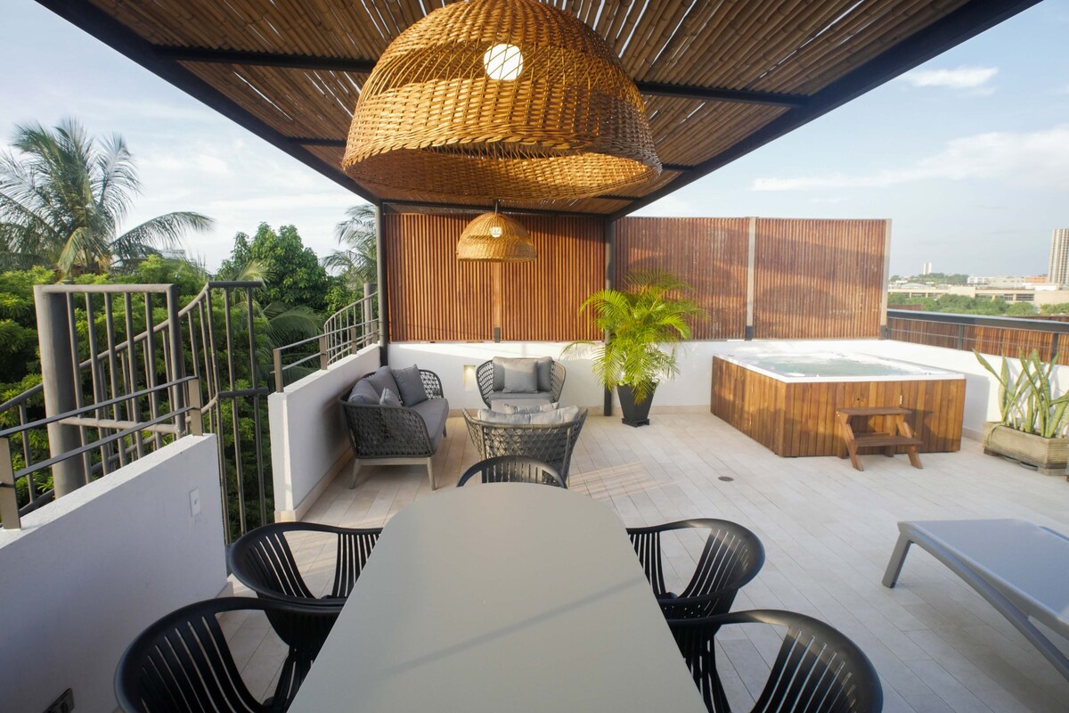 A private rooftop terrace features a dining table surrounded by comfortable chairs, with a lounge area consisting of two armchairs and a coffee table. A jacuzzi is positioned to the side, and a pergola provides shade, enhancing the view of surrounding greenery.