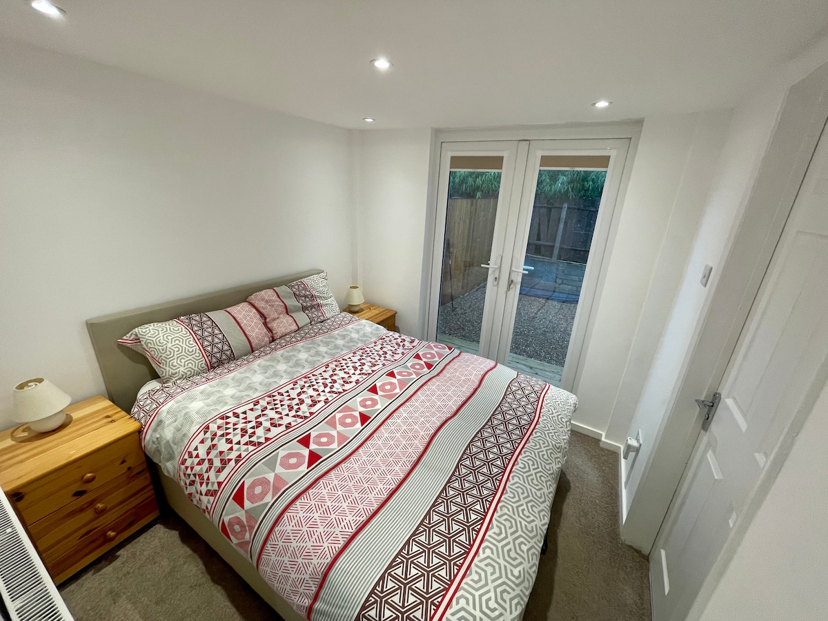 A double bedroom is shown, featuring a spacious bed with a patterned duvet and two nightstands on either side. Natural light enters through double patio doors, which lead to the garden. The walls are painted white, complementing the soft carpet.