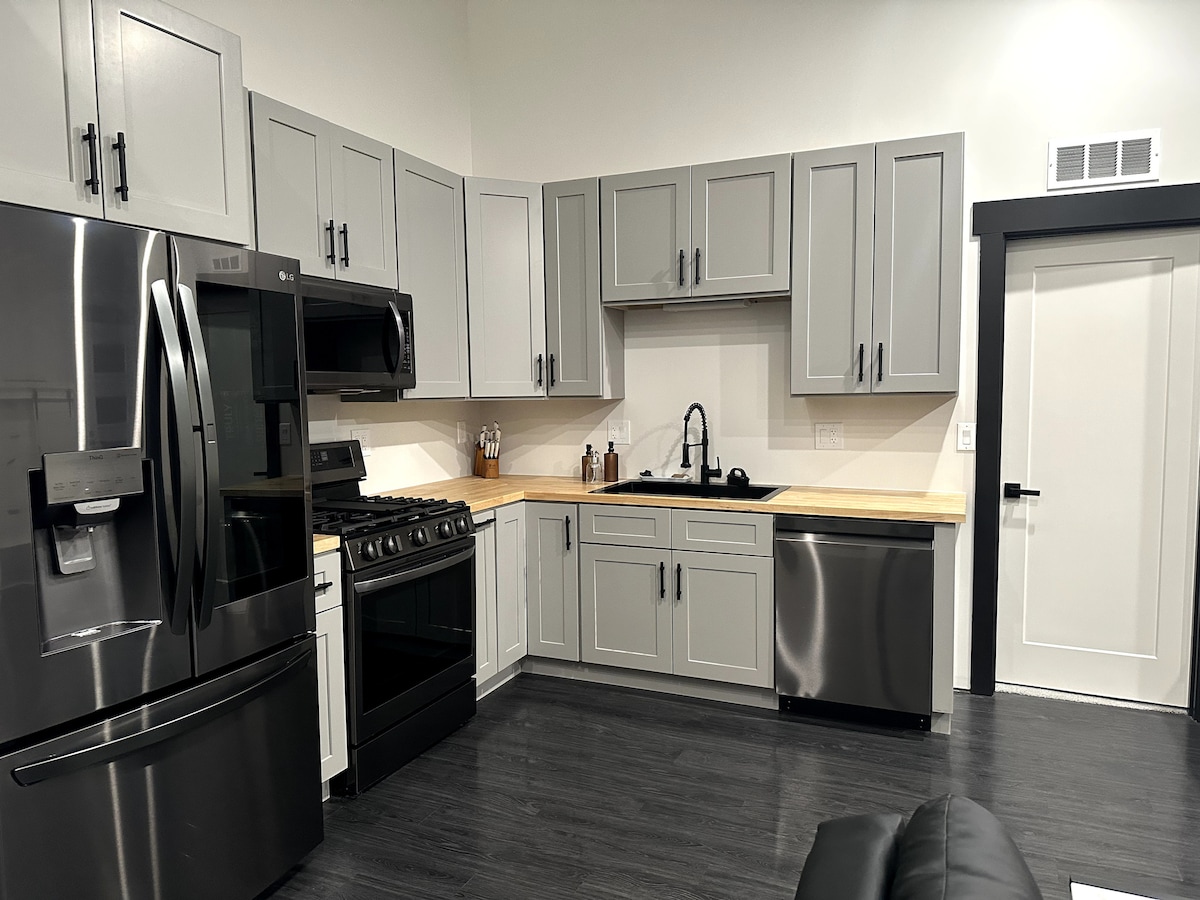 A modern kitchen is equipped with stainless steel appliances, including a refrigerator, stove, and dishwasher. Light gray cabinets line the walls, complemented by a wooden countertop. A sleek black sink is situated beneath the window, which is positioned near a door leading to the next area.
