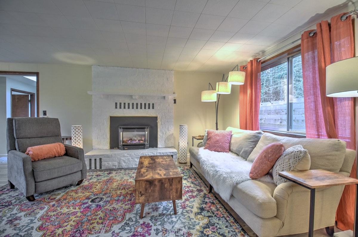 A cozy living room features a light-colored sofa with a soft throw, complemented by an accent chair. A wooden coffee table rests on a colorful area rug. The gas fireplace is set within a white stone wall, while large windows allow natural light, adorned with sheer curtains.