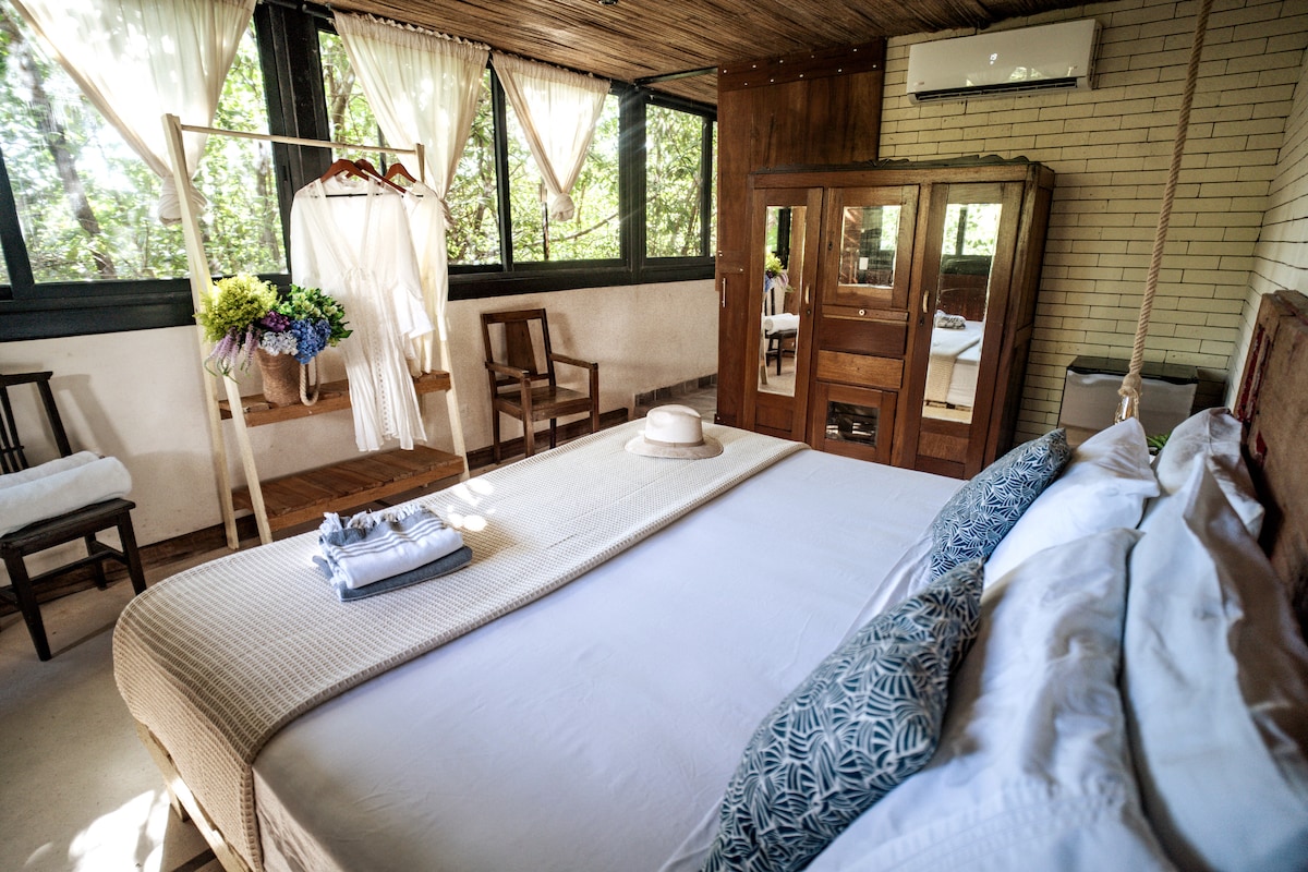 A spacious room is highlighted by a king-size bed, featuring crisp white linens and decorative pillows. Natural light filters through large windows adorned with sheer curtains. Nearby, a wooden wardrobe and two chairs provide additional furnishings, while a hat rests on the bed for a touch of charm.