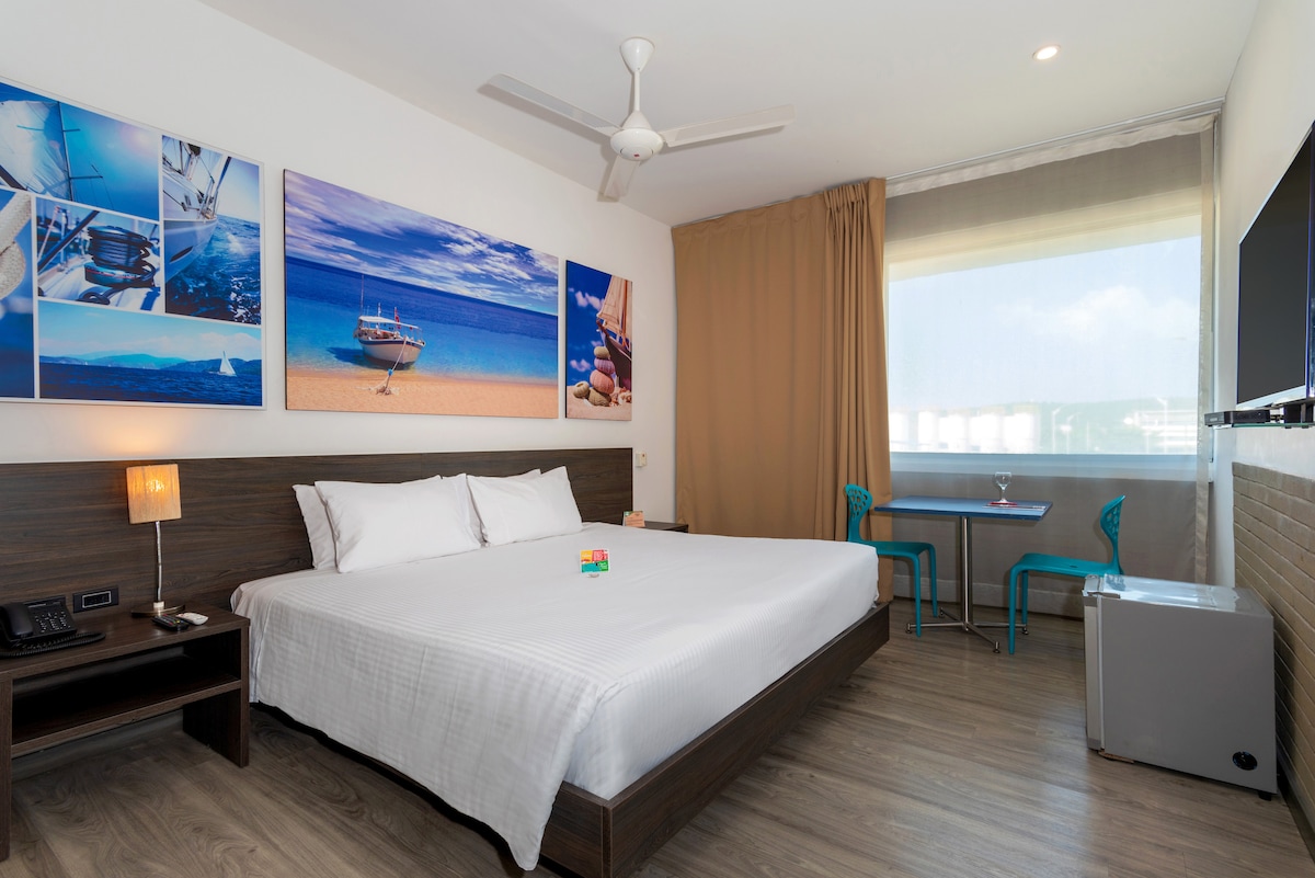 A spacious bedroom features a large bed with crisp white linens and a minimalist design. Colorful artwork depicting serene beach scenes adorns the wall above the bed. Natural light enters through a large window, illuminating a small table and chairs in the corner, alongside a mini fridge.