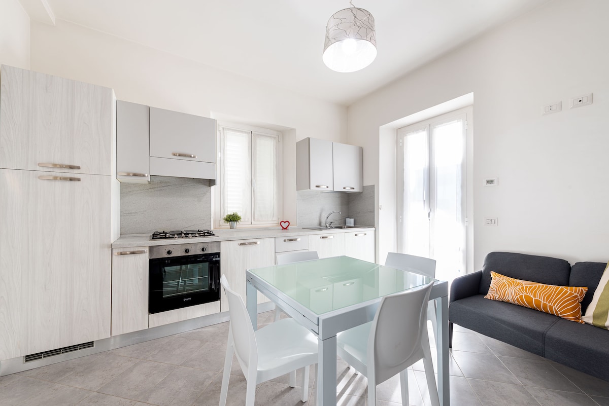 A modern kitchen area features light cabinetry and a sleek grey countertop. A glass dining table is surrounded by four white chairs. A sofa with stripes sits nearby, and light pours in through a large window, creating a bright and airy environment.