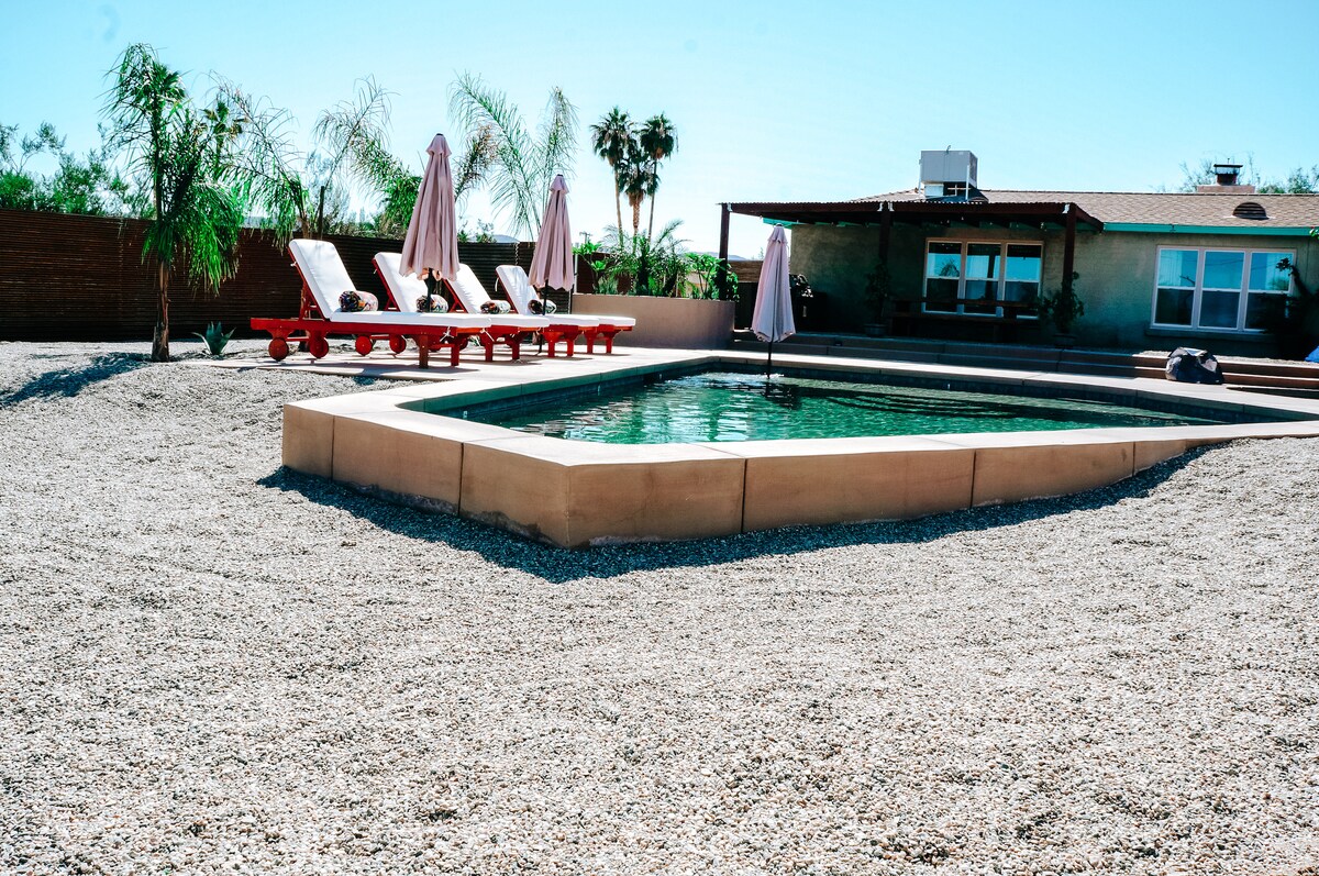 A spacious swimming pool is surrounded by a gravel area, featuring several lounge chairs under large umbrellas. In the background, the home is visible with multiple windows and a covered patio, enhancing the outdoor retreat ambiance.