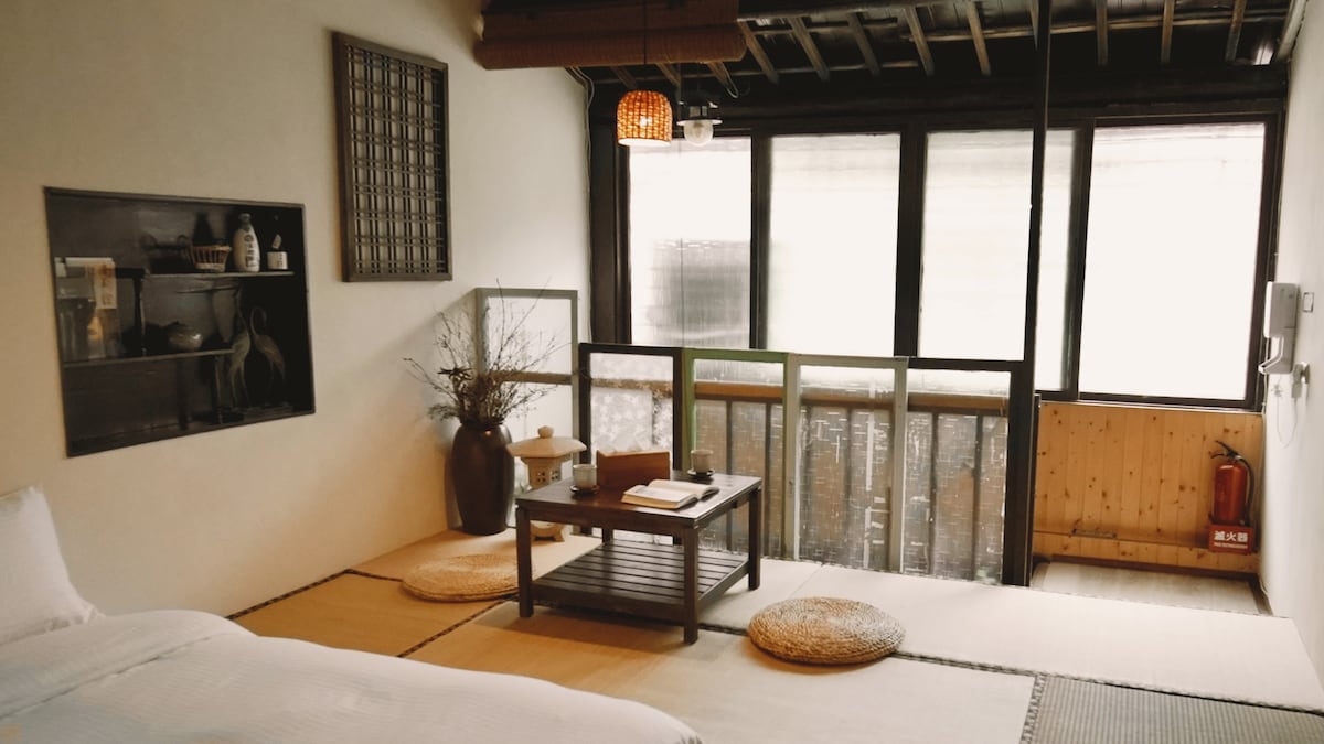 A serene room featuring tatami flooring and large windows that allow natural light to fill the space. A low wooden table is positioned in the center, surrounded by round cushions. A minimalist display shelf is visible, showcasing various items for ambiance.