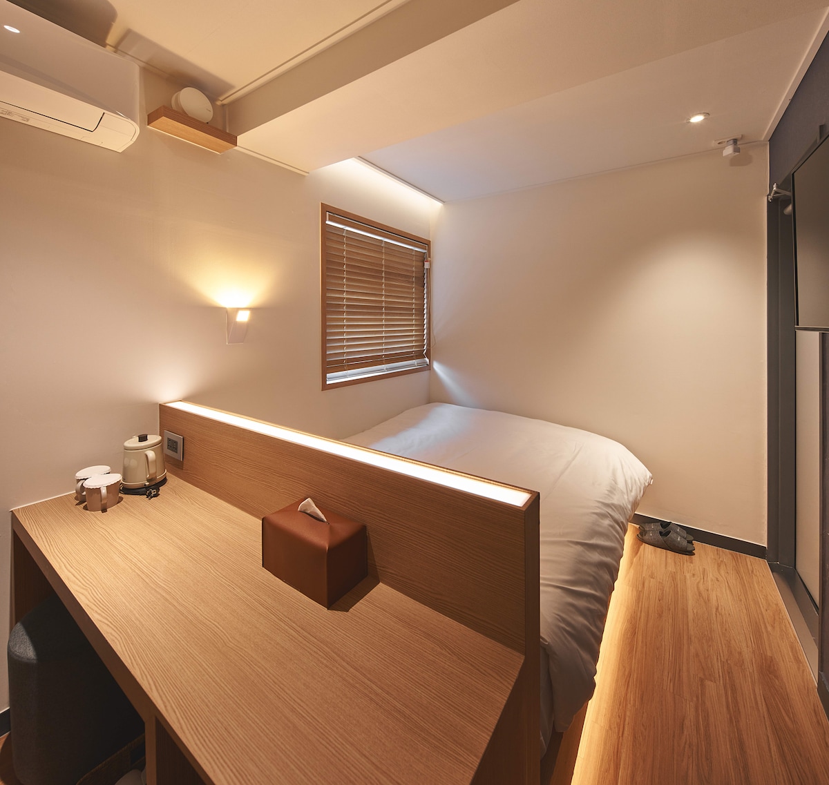 A warm-toned room features a spacious bed against a wooden headboard. A desk with a seating area is positioned nearby, complemented by a small lamp and an electric kettle. Natural light filters through a window adorned with wooden blinds, enhancing the room's quiet ambiance.