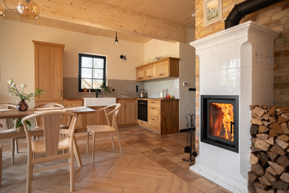 A spacious kitchen and dining area features warm wooden cabinetry and a modern dining table with four chairs. A stove with firewood stacked nearby adds a cozy element, while natural light enters through a large window framed in a neutral wall.