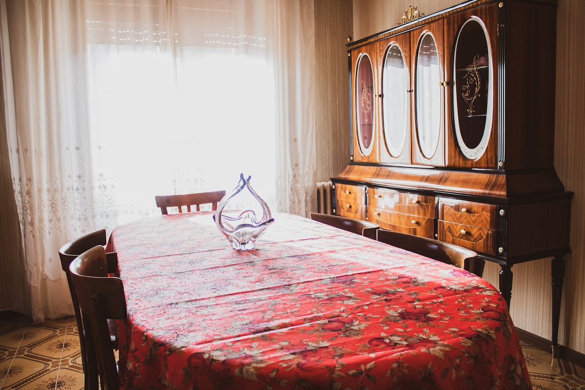A dining area is presented with a large rectangular table topped with a red floral tablecloth. Surrounding wooden chairs complement a vintage wooden cabinet with glass panels, showcasing decorative items. Natural light enters through sheer curtains, softening the space.