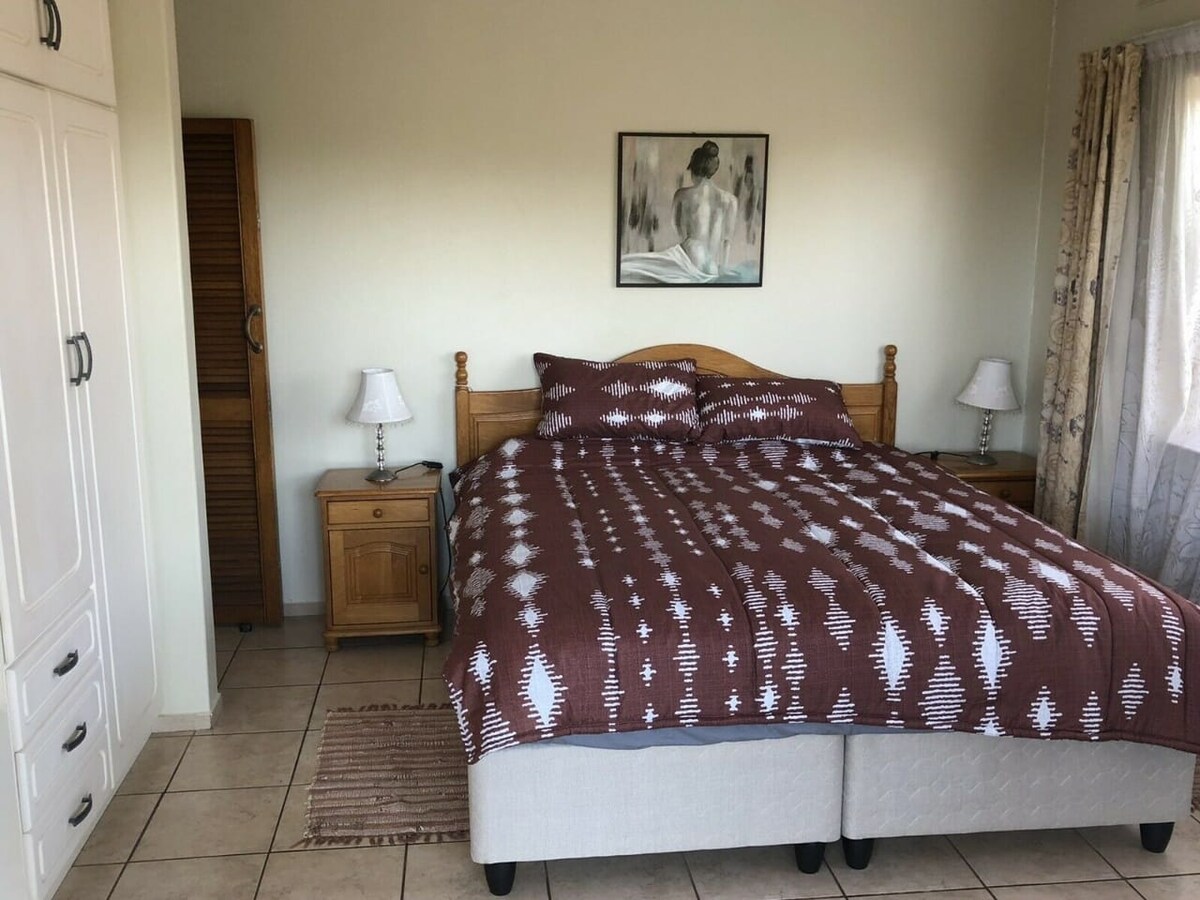 A spacious bedroom features a large bed dressed in a patterned brown and white coverlet. Bedside tables with lamps are positioned on either side, and a piece of art adorns the wall above the bed. Natural light filters through a window dressed with light curtains.