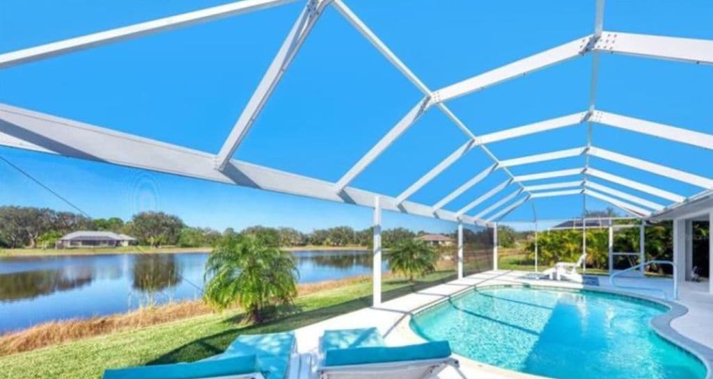 A spacious pool area is showcased, featuring a clear blue pool surrounded by a screened-in enclosure. Lush greenery and a serene lake are visible in the background, providing a tranquil setting. The bright sky enhances the open atmosphere of the space.