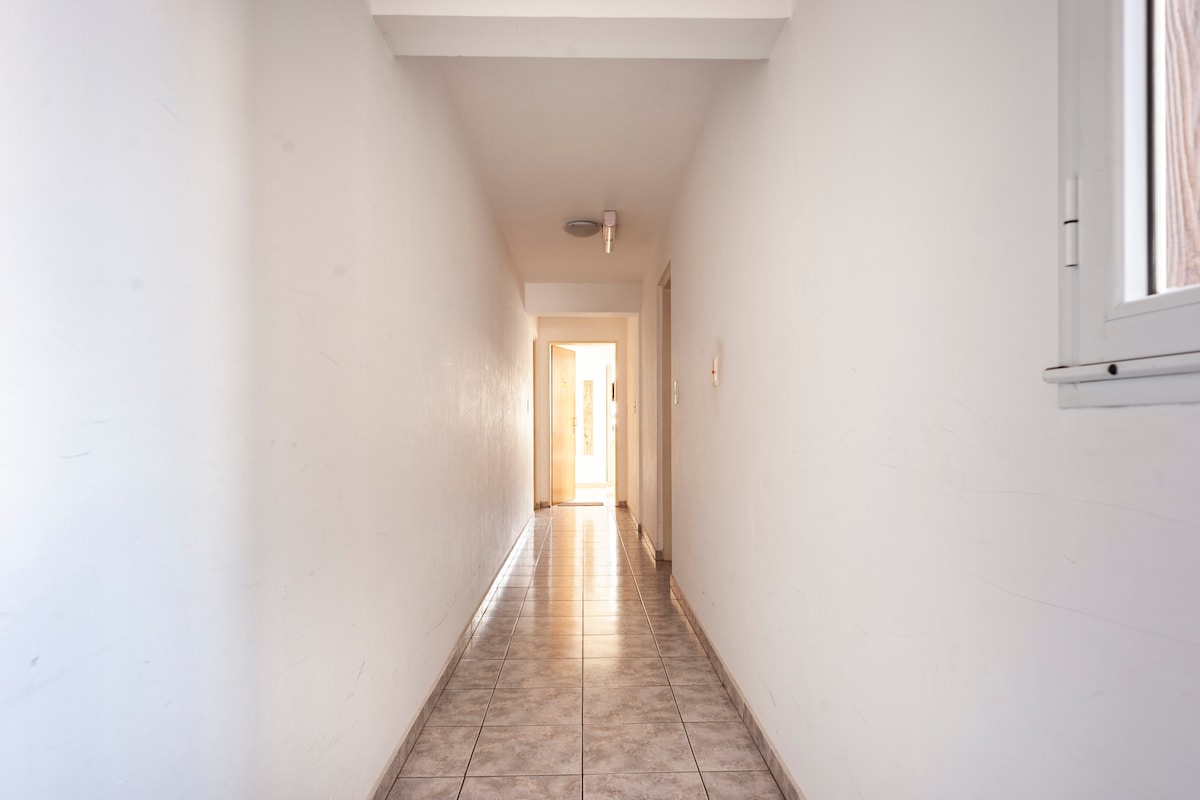 A long, narrow hallway features smooth tile flooring that reflects natural light. The walls are painted in a clean white, and natural light is visible at the end of the corridor, providing a sense of brightness and openness.
