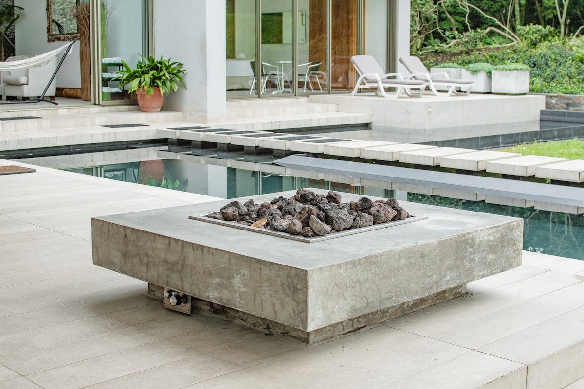 A modern firepit made of concrete, positioned on a stone patio. Surrounding the firepit is a serene area with a tranquil swimming pool and lounge chairs visible in the background, bordered by landscaped greenery.