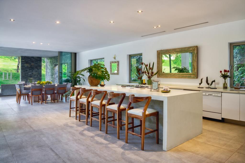 An open kitchen space features a large white island with seating for eight. Natural light filters through large windows, illuminating the area. It is connected to a dining table that seats twelve, creating an inviting atmosphere for communal meals.