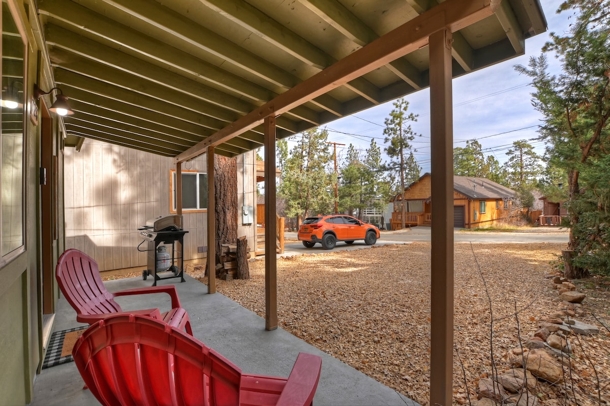 A covered porch is showcased, featuring two red Adirondack chairs positioned for outdoor relaxation. A charcoal grill is visible next to the seating area. The gravel driveway leads to the street, where a parked orange car can be seen amid the surrounding trees.