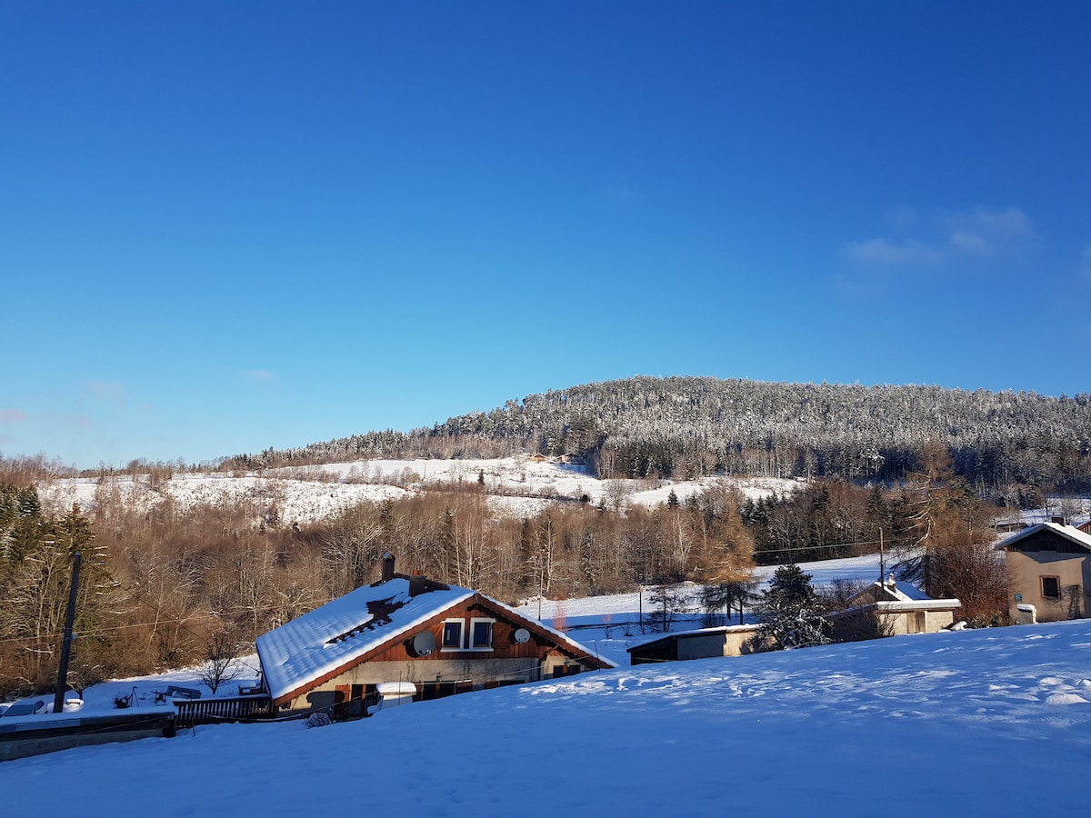 A serene winter landscape showcases snow-covered rolling hills and a clear blue sky. The view includes charming wooden structures nestled among the trees, highlighting the tranquil retreat environment. The undisturbed snow blankets the ground, creating a peaceful outdoor setting.