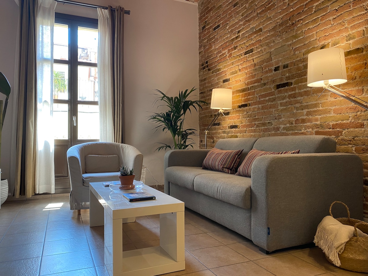 The living area features a cozy gray sofa and a white coffee table, complemented by a comfortable armchair. A decorative plant adds a touch of greenery. Natural light streams in through the large windows, illuminating the exposed brick wall and providing a warm ambiance.