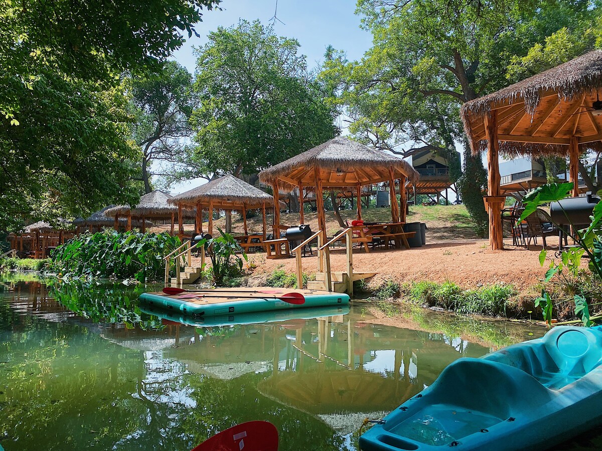 A serene view of shaded cabanas lined along the creek, surrounded by lush greenery. Two kayaks are moored at the water's edge, reflecting the vibrant trees above and the inviting surface of the water.