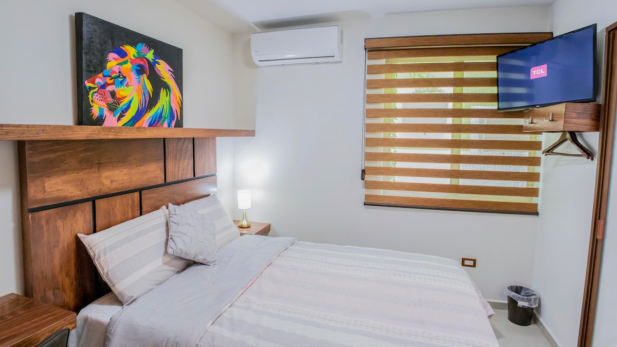 A cozy bedroom features a spacious bed adorned with light-colored linens. A colorful lion artwork is displayed above the headboard, while a flat-screen TV is mounted on the wall. Ambient light is provided by a bedside lamp, and transparent window treatments allow soft light to filter in.