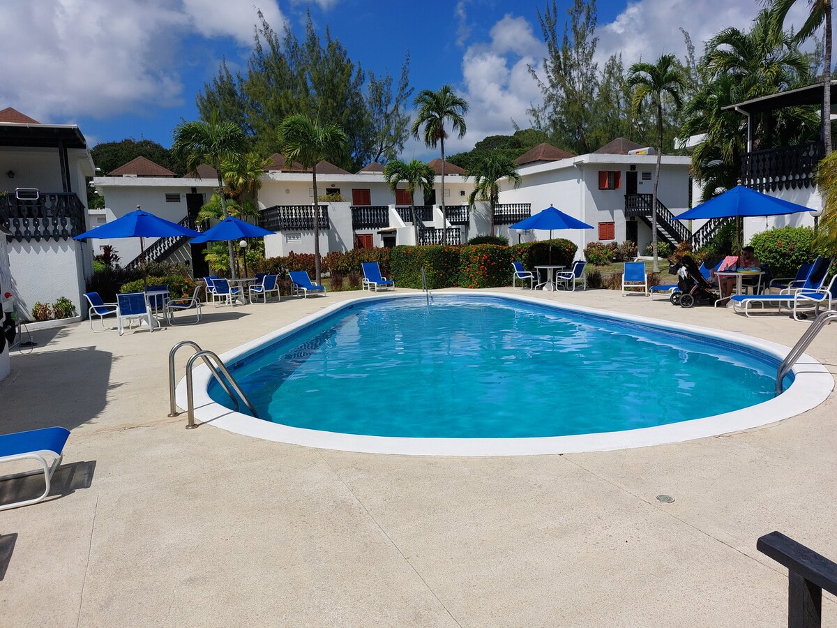 A blue swimming pool is surrounded by lounge chairs with umbrellas, offering shaded seating. Lush greenery and well-maintained landscaping enhance the area's inviting feel, while the buildings of the resort are visible in the background under a clear blue sky.