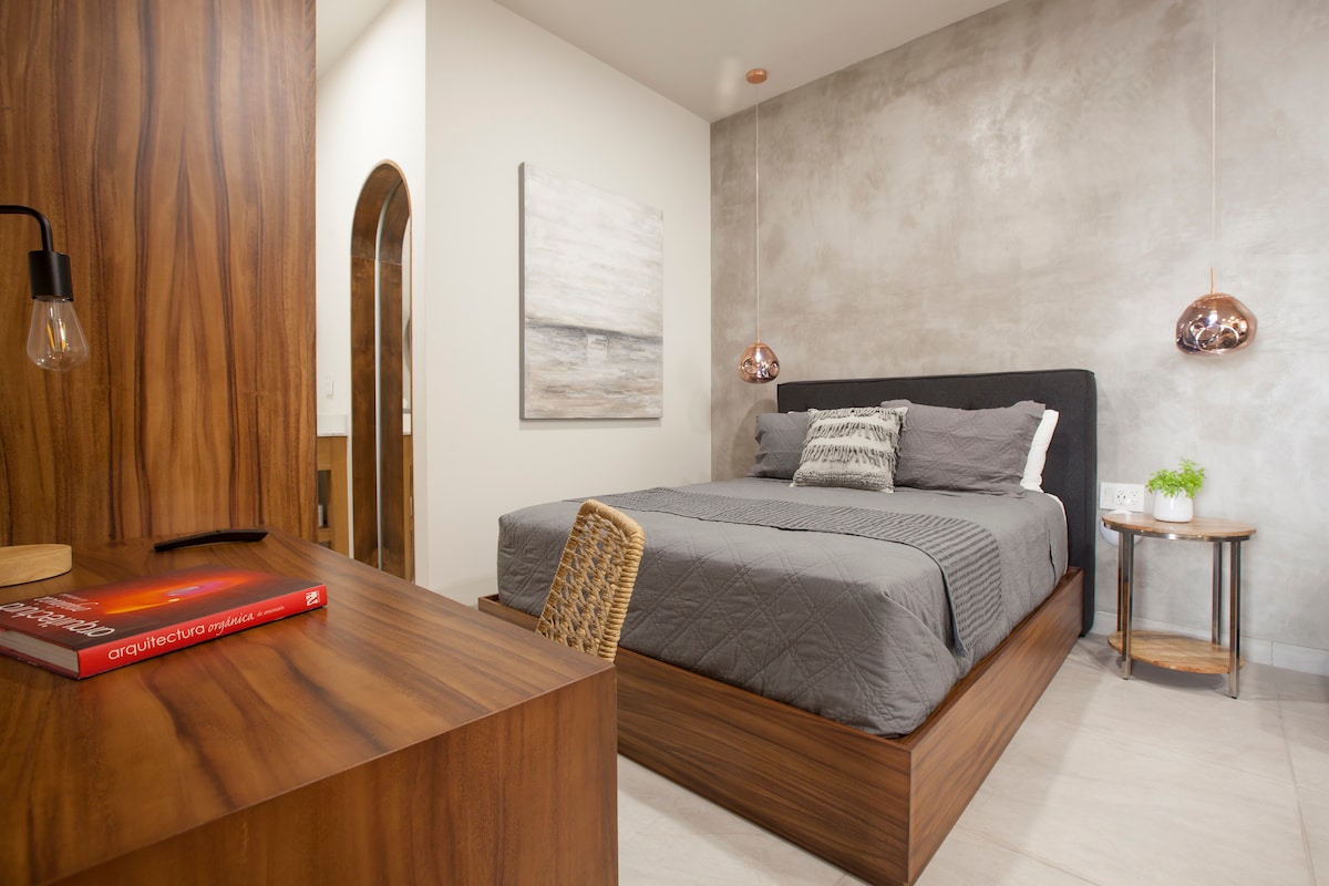 A modern sleeping area features a spacious bed with gray bedding, flanked by warm pendant lighting. A simple desk with a woven chair and a small table with a plant highlight the natural wood tones, while a textured wall art adds a touch of sophistication.