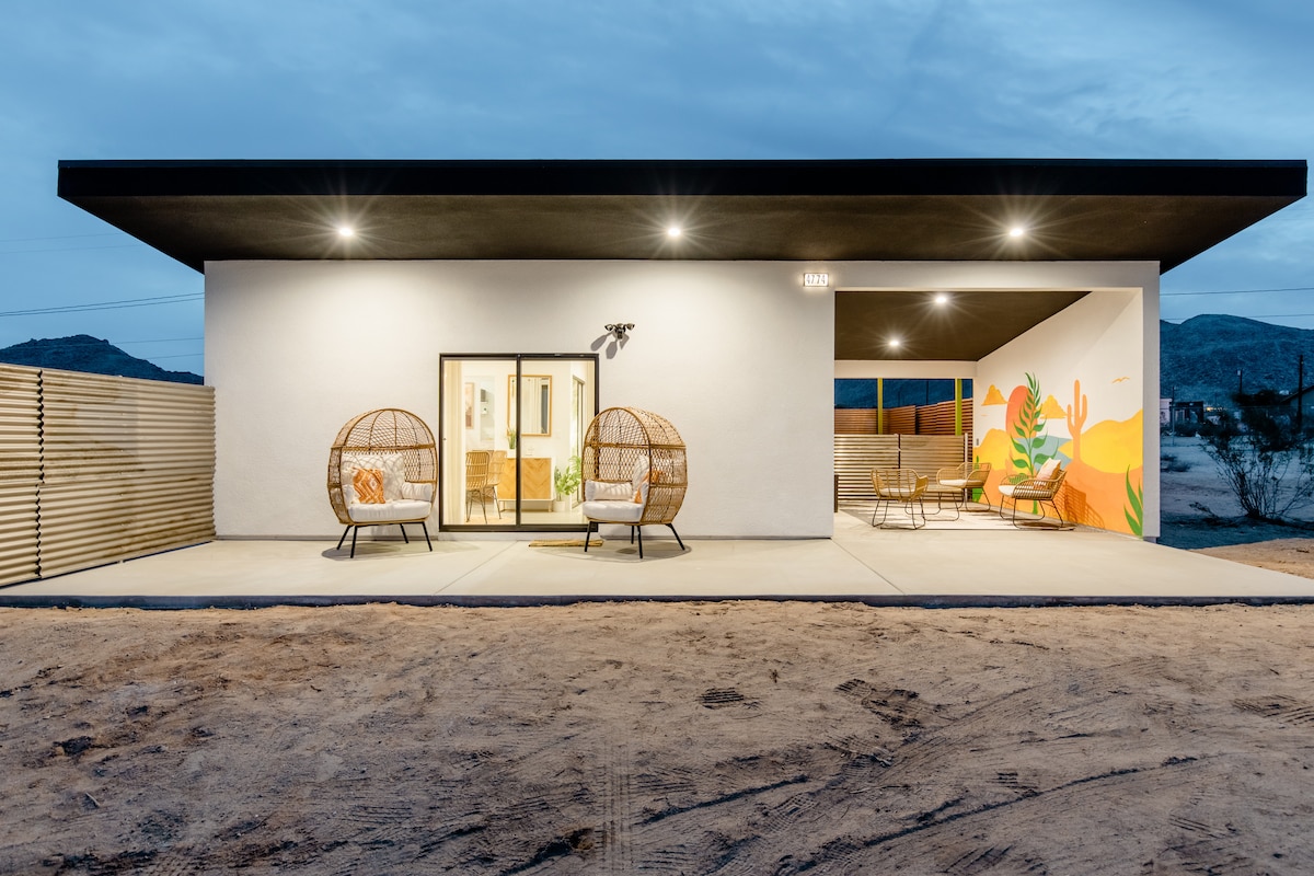 A modern exterior of the tiny home showcases a smooth, white facade complemented by a black roof. Two woven hanging chairs are positioned in front of large glass doors, providing an inviting entrance. A vibrant mural adds a splash of color to the side wall.