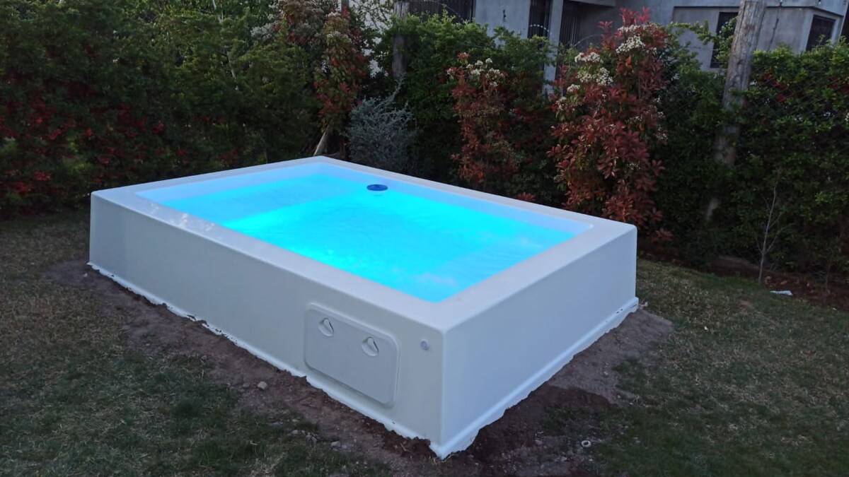 A small swimming pool is situated in a lush garden, with vibrant greenery and flowering shrubs surrounding it. The pool is designed with a clean, modern edge and is illuminated with soft blue lighting, creating a serene atmosphere in the outdoor space.