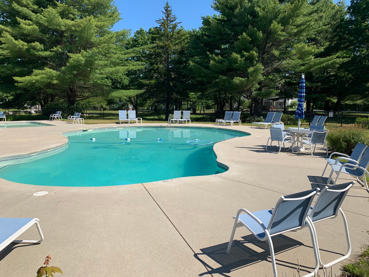 A sparkling outdoor pool is surrounded by lounge chairs on a sunlit patio. Lush green trees provide shade in the background, and blue umbrellas are positioned nearby. Clear water reflects the sunlight, creating a refreshing atmosphere.