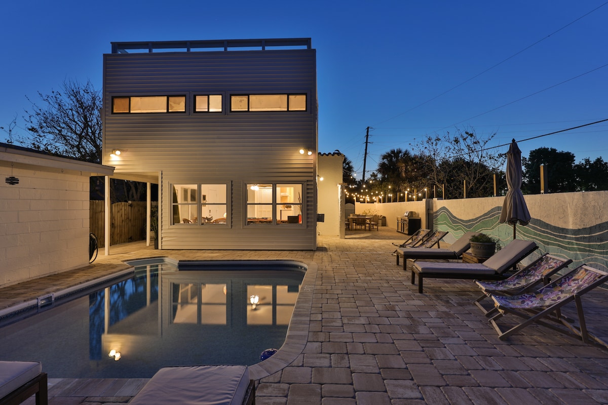The outdoor area features a small swimming pool surrounded by a deck with lounge chairs. A two-story structure with large windows is seen in the background, illuminated by warm lighting. String lights create a cozy ambiance over the poolside area.