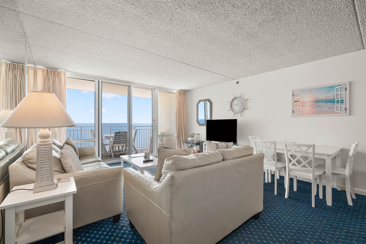The living area is furnished with a light-colored sofa and armchairs, complemented by a white coffee table. A flat-screen TV is positioned near a dining table with seating for four. Views of the ocean can be seen through expansive glass doors, leading to a private balcony.