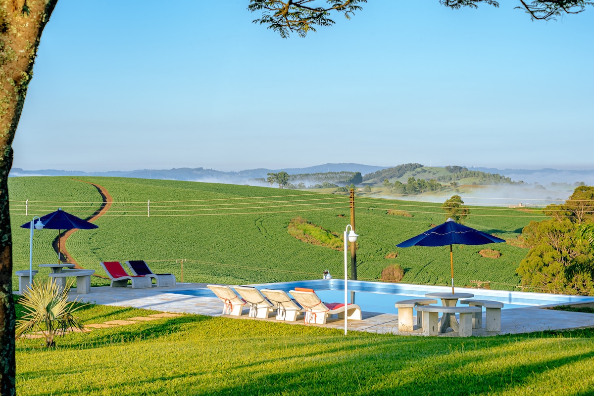 A panoramic view reveals a tranquil pool surrounded by lounge chairs and umbrellas. Lush green fields stretch into the distance, framed by gentle hills, while a winding dirt path bisects the landscape under a clear blue sky.