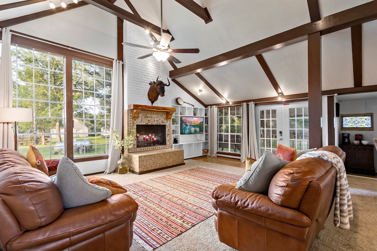The living room features a spacious layout with high, exposed beam ceilings. A large window allows natural light to fill the space, highlighting the cozy leather sofas and textured area rug. A brick fireplace serves as a focal point, complemented by a mounted flat-screen TV.