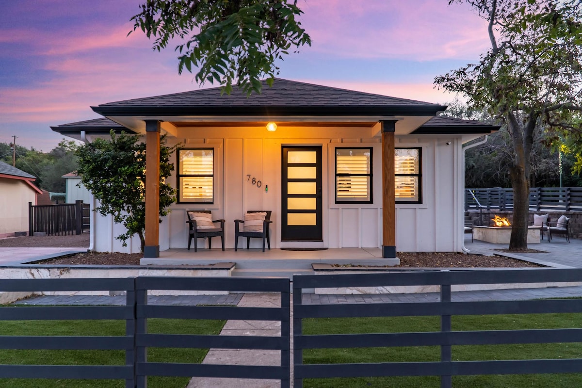 A charming home features a welcoming entry with a distinct front door and warm lighting. Two outdoor chairs are positioned on the porch, surrounded by landscaped greenery. The pathway leads to a fenced yard, with a visible outdoor seating area in the background.
