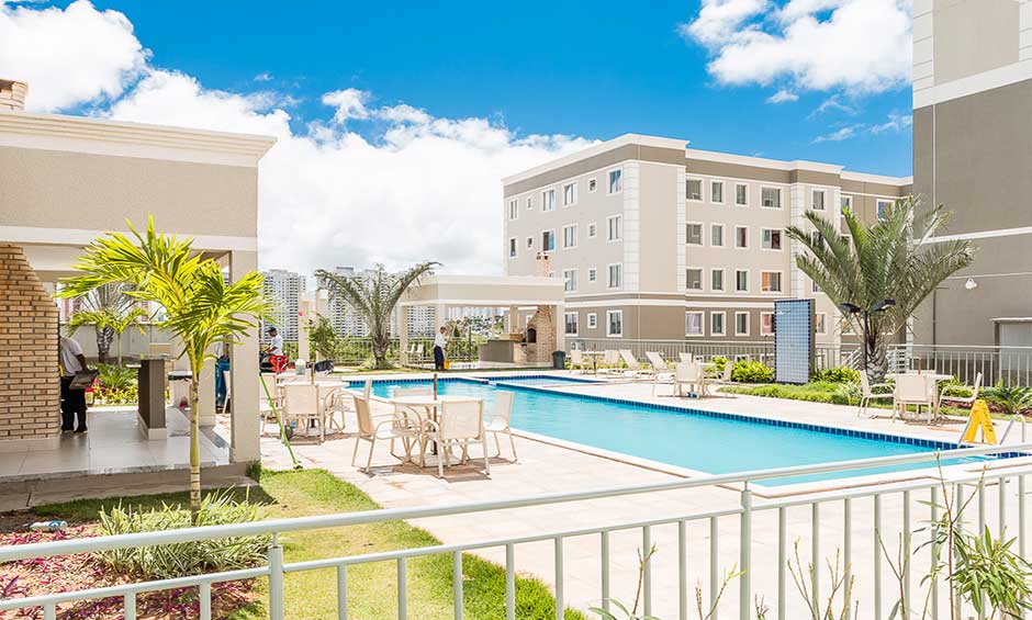 A clear view of the outdoor pool area is presented, surrounded by lounge chairs and vibrant greenery. The modern buildings in the background provide a backdrop, and palm trees frame the scene under a bright blue sky, creating a welcoming atmosphere.