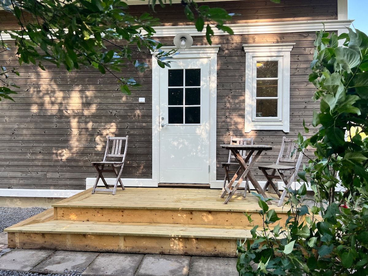 A wooden deck features two folding chairs and a small table, providing an outdoor seating area. The entrance of the two-story structure is adorned with a white door and a side window, framed by greenery that adds a natural touch.