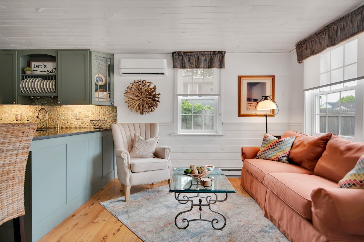 A cozy living area is highlighted by a comfortable peach-colored sofa adorned with colorful cushions. A glass coffee table sits at the center, complemented by a stylish armchair. Natural light enters through two windows, enhancing the quaint decor and warm ambiance.