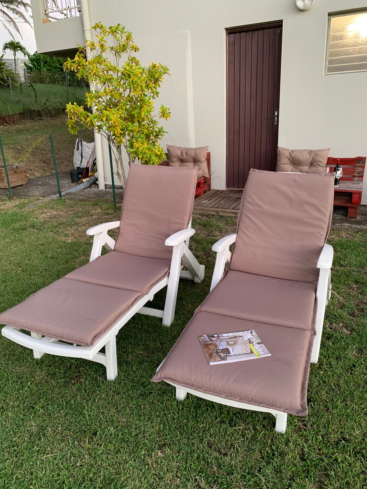 Two comfortable lounge chairs are positioned on a grassy area, featuring plush cushions and a small side table with a magazine. A modest wooden door is visible in the background, along with a few outdoor seating options, creating a welcoming space for relaxation.