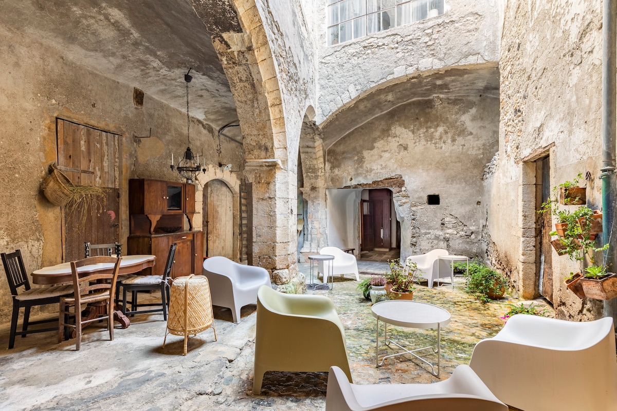 An open courtyard is surrounded by weathered stone walls, displaying high arches and exposed brick. Seating arrangements include simple white chairs and a round table, complemented by greenery and potted plants. Natural light filters through a large window, highlighting the tranquil outdoor space.