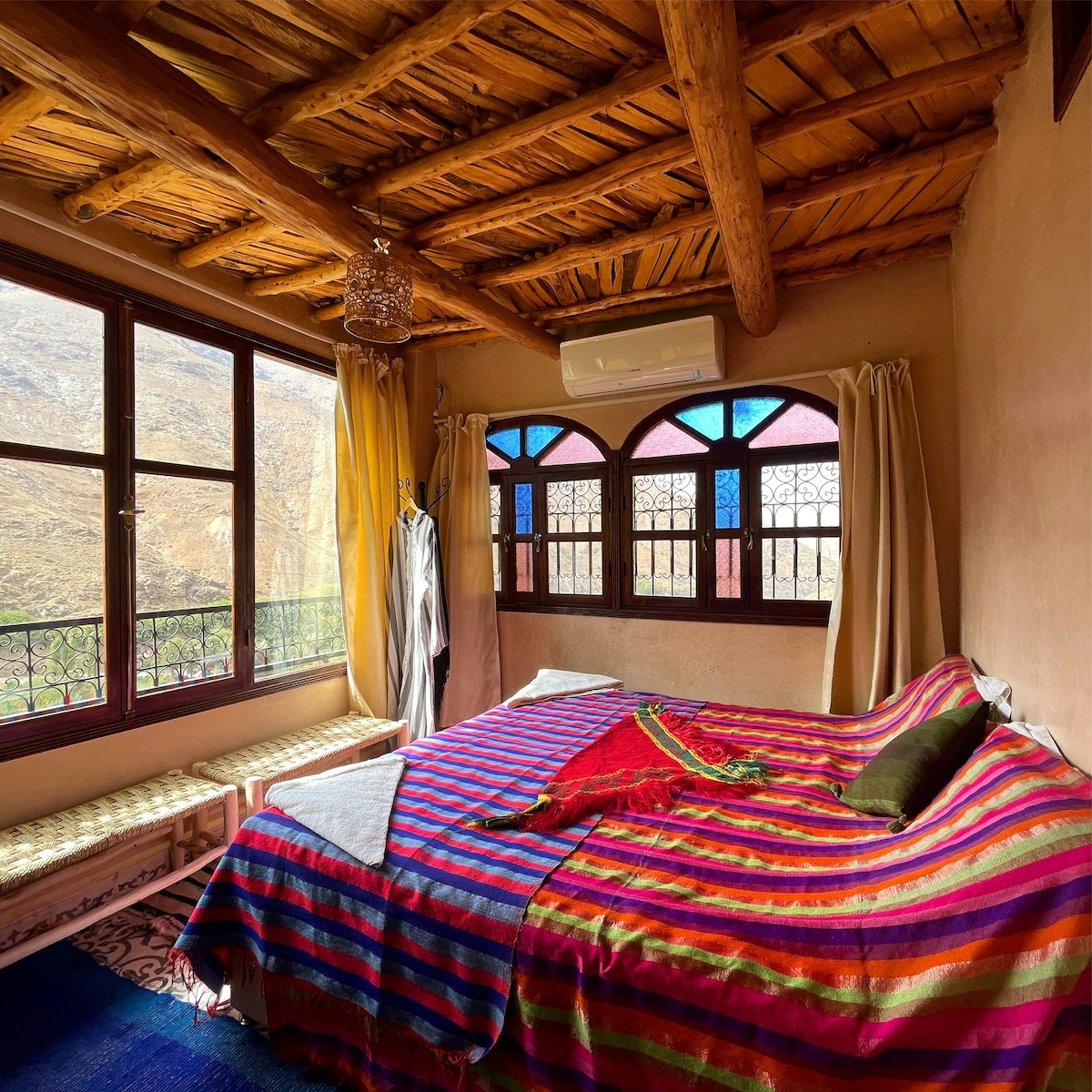 A comfortable bedroom features a large bed with colorful striped bedding. Warm wooden beams are visible on the ceiling, while large windows adorned with decorative panes offer scenic mountain views. Curtains softly frame the windows, creating a bright and inviting atmosphere.