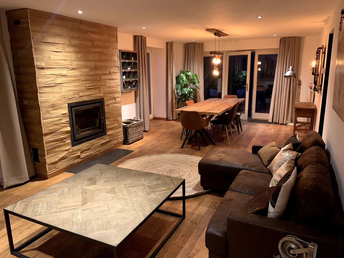 A spacious living area features a large brown sectional sofa and a textured coffee table on a natural fiber rug. A rustic dining table accommodates several chairs, while a stone fireplace adds a warm touch. Natural light filters in through large doors, framed by neutral curtains.