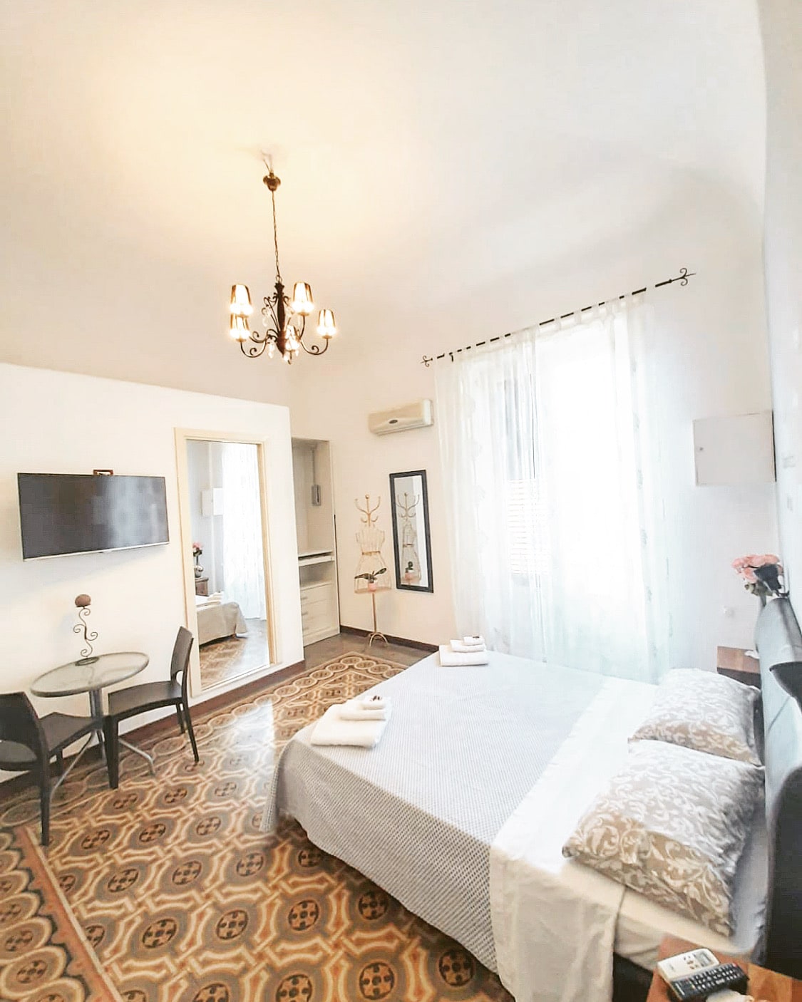 A well-lit bedroom features a spacious bed covered with soft linens and decorative pillows. A small round table with two chairs is positioned nearby. A flat-screen TV is mounted on the wall. Light pours in through sheer curtains, enhancing the room's airy feel.