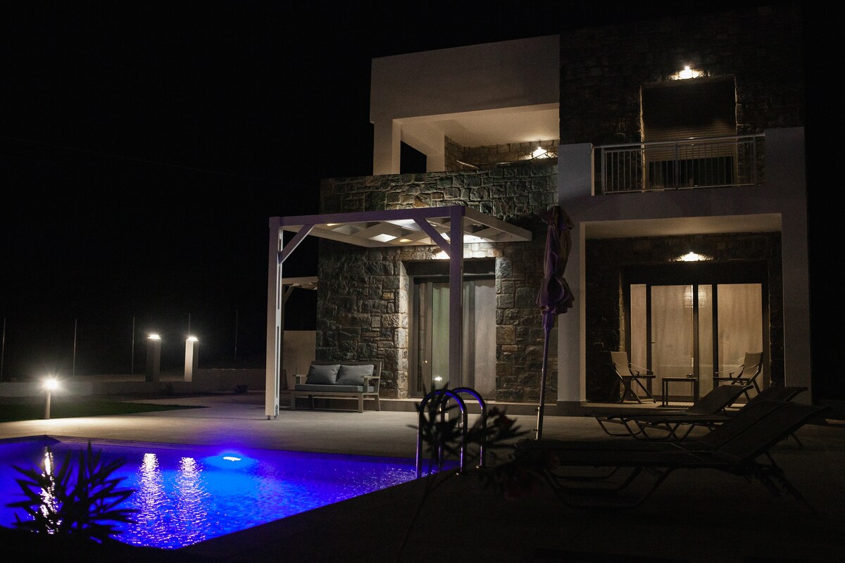 The modern apartment is illuminated with soft lighting, showcasing a private swimming pool featuring subtle blue hues. A spacious terrace is visible with lounge chairs and a shaded seating area. The building's stone façade stands out against the night sky, providing a tranquil ambiance.