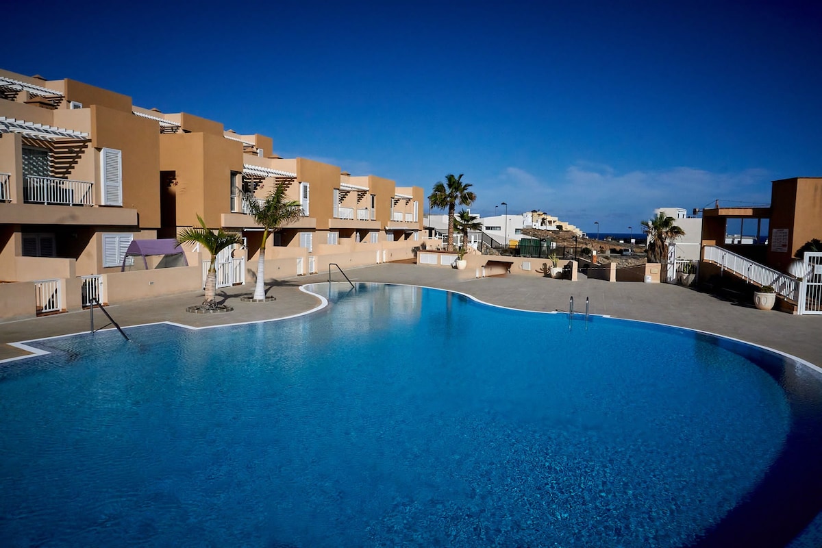 A large outdoor pool is surrounded by spacious terraces and palm trees. The clear blue water reflects the sunny sky, creating a tranquil atmosphere. Apartment units are visible in the background, showcasing neutral tones and balconies, contributing to the overall inviting setting.