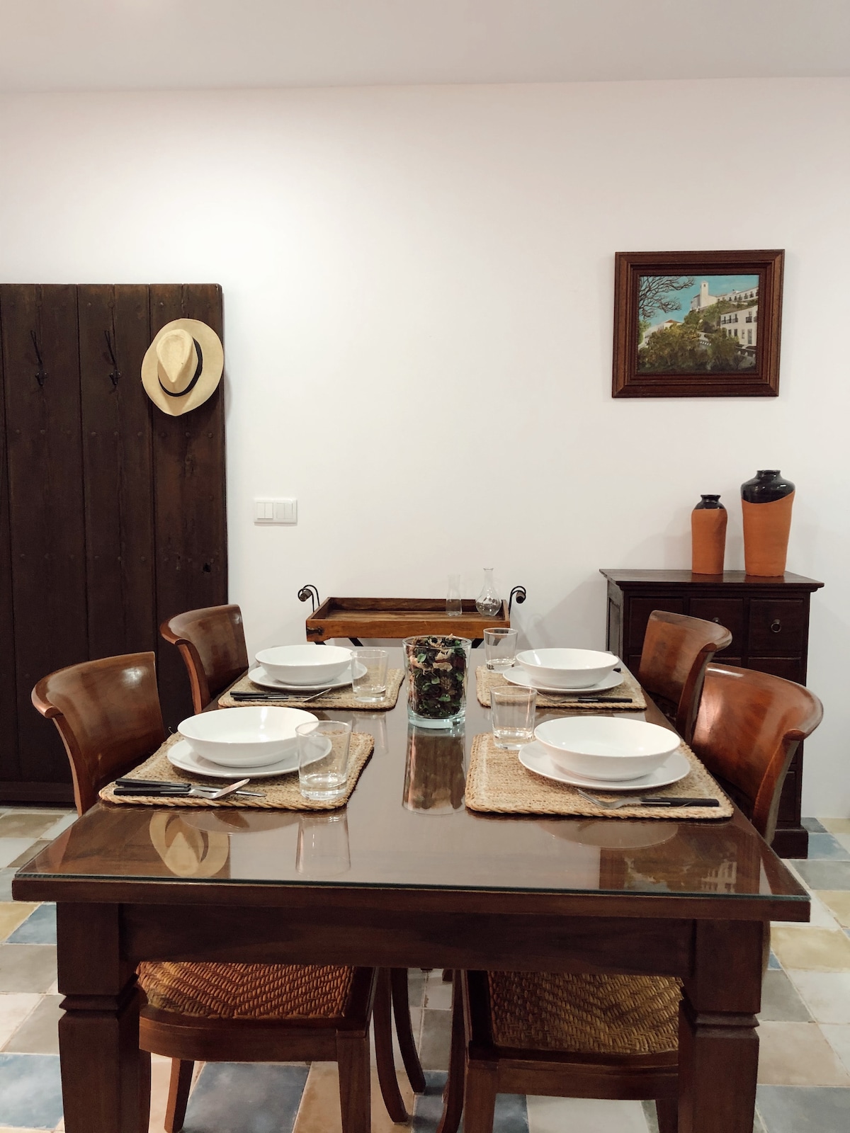 A dining area is presented with a polished wooden table and six woven chairs. The table is set with white plates and glasses. A decorative vase is centered on the table, while a framed painting hangs on the wall next to a rustic wooden cabinet, accented by a stylish hat.