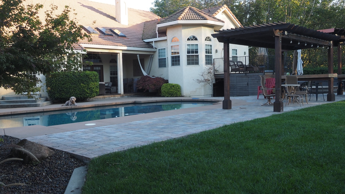 A landscaped backyard features a rectangular swimming pool surrounded by a stone patio. A shaded pergola provides seating, while lounge chairs are positioned nearby. The house is visible in the background, showcasing large windows and a warm exterior.