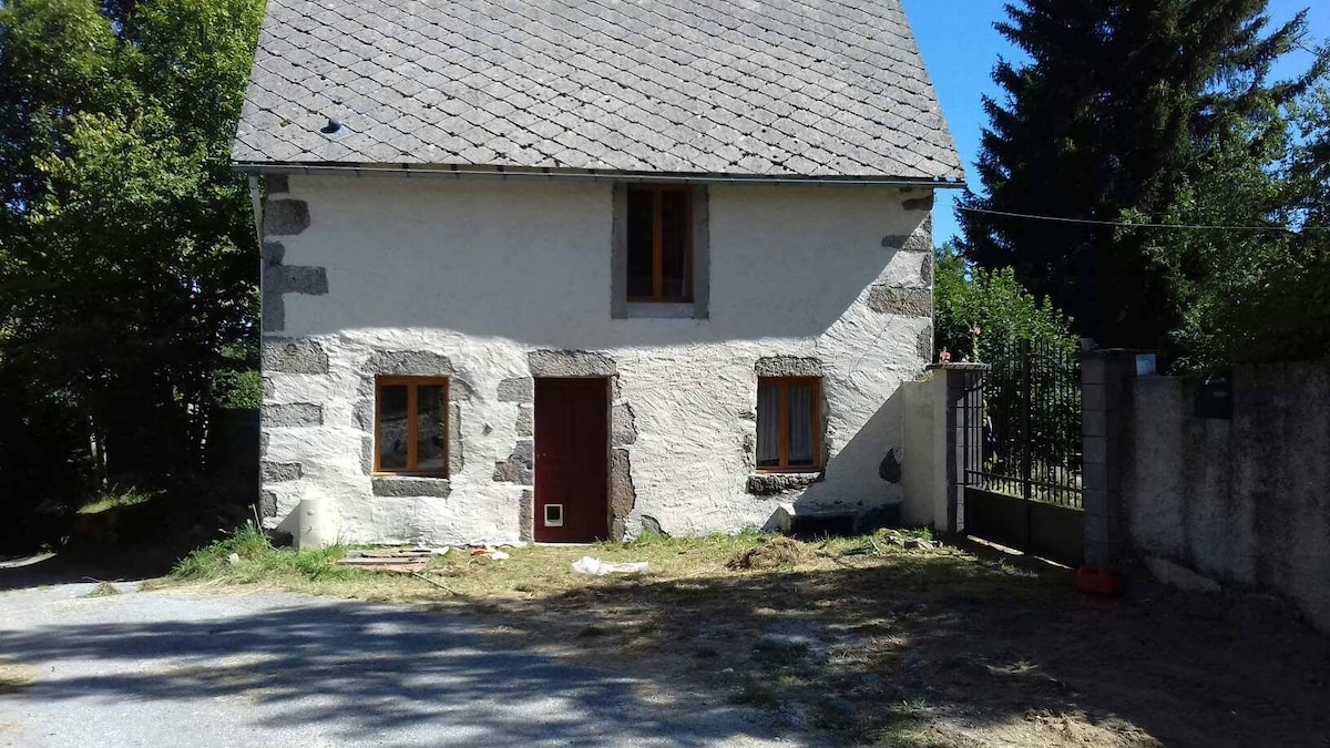 A two-story house exhibits a rustic charm with a stone facade and wooden windows. The entrance features a red door, while the roof is covered with traditional slate tiles. Surrounding greenery adds to the peaceful setting, creating an inviting outdoor space.